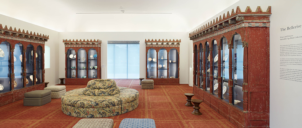 Four wooden showcases with artifacts inside along the back three walls of the room, red carpet and large circular couch seat in the middle of the room.